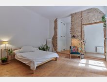 Schlafzimmer mit minimalistischer Einrichtung, bestehend aus einem Holzrahmenbett mit weißer Bettwäsche, Pflanzen auf einem kleinen Regal, einer Stehleuchte und einem Sessel. Eine Wand aus freigelegtem Ziegelstein mit einem Durchgang sowie Holzböden und weiße Wände verleihen dem Raum einen rustikalen Charme.