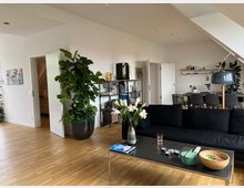 Wohnzimmer in einer modernen Wohnung mit Holzfußboden, einer schwarzen Couch und einem rechteckigen Couchtisch, auf dem verschiedene Gegenstände wie eine Blumenvase, Schalen und Fernbedienungen stehen. Im Hintergrund befinden sich eine offene Küche mit Esstisch, ein Regal und große grüne Zimmerpflanzen.