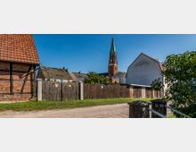 Eine ländliche Szene mit Fachwerkhäusern und Ziegeldächern, einem Holzzaun im Vordergrund und einem Kirchturm aus rotem Backstein mit grünem Spitzdach im Hintergrund. Der Himmel ist klar und blau, und es sind kleine Bäume und Sträucher zu sehen.
