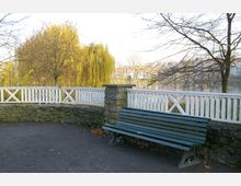 Eine Parkszene mit einer grünen Holzbank im Vordergrund, umgeben von einem niedrigen Steinrand und einem weißen Holzgeländer. Im Hintergrund sind herbstliche Bäume, ein Fluss und vereinzelte Gebäude sichtbar.
