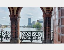 Blick durch die roten Backsteinbögen der Oberbaumbrücke in Berlin auf die Spree, mit modernen Hochhäusern im Hintergrund und dekorativen schmiedeeisernen Geländern im Vordergrund. rechts und links sind Gebäude entlang des Flusses zu sehen.