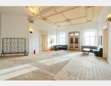 Großzügiger, heller Innenraum mit weiß verputzten Wänden, hohen Fenstern und einer doppelflügeligen Eingangstür. Auffällig sind die dekorierte Decke mit Kronleuchter, der abgenutzte Fliesenboden sowie Sitzgruppen mit grünen Sofas und kleinen Tischen entlang der Wände.