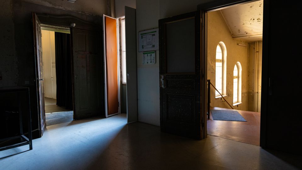 Dunkler Innenflur eines älteren Gebäudes mit mehreren offenen Türen, durch die Tageslicht in den Raum fällt. Rechts ist ein Treppenhaus mit hohen, bogenförmigen Fenstern und abgenutzten Wänden zu sehen.