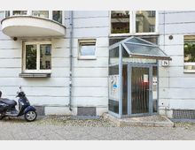 Mehrfamilienhaus mit heller Fassade, einem verglasten Eingangsbereich mit Flachdachvordach und Hausnummer 74. Links steht ein Motorroller geparkt auf einem Gehweg mit Kopfsteinpflaster.