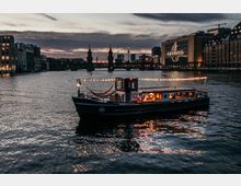 Ein beleuchtetes Boot mit Lichterkette fährt auf einem Fluss bei Dämmerung, im Hintergrund die Oberbaumbrücke in Berlin sowie umliegende Gebäude, darunter das Universal-Gebäude mit großem beleuchtetem Schriftzug.