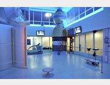 Innenraum eines modernen Raumfahrt-Trainingszentrums mit einer detailgetreuen Nachbildung eines sowjetischen Sojus-Raumfahrzeugs, umgeben von Trainingsgeräten wie einem Laufband. Der Raum ist von Glaswänden umgeben, die in blauem Licht beleuchtet sind, und es hängen mehrere Bildschirme an den Wänden.