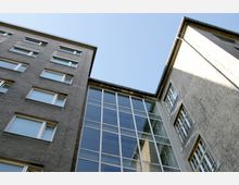 Blick von unten auf mehrere graue Gebäude mit rechteckigen Fenstern, verbunden durch eine Fassade mit großflächigen Glasfenstern. Der Himmel ist klar und blau im Hintergrund.