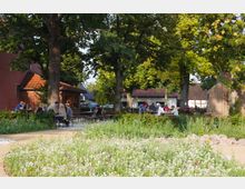 Biergarten im Freien mit Holzbänken und -tischen, umgeben von hohen Bäumen und einer blühenden Blumenwiese im Vordergrund. Im Hintergrund sind Gebäude mit roten Ziegeldächern und Fachwerkelementen zu sehen.