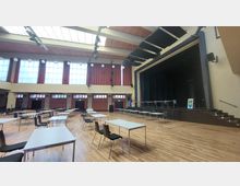 Ein großer Veranstaltungssaal mit Holzboden, einer erhöhten Bühne auf der rechten Seite und mehreren weißen Tischen mit schwarzen Stühlen im Raum verteilt. Die Fensterfront im Hintergrund besteht aus großen, quadratischen Glaselementen, teils mit roten Vorhängen.