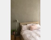 Schlafzimmer mit einem beigen Polsterbett, weißer Bettwäsche und einem rosa Zierkissen. Links an der Wand hängt eine kleine weiße Lampe, darunter steht ein Holzhocker mit einer Topfpflanze.