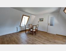 Ein schlichtes Zimmer mit weiß gestrichenen Wänden, einem schrägen Dachfenster mit Holzrahmen, Holzboden und einem kleinen runden Tisch mit vier gepolsterten Stühlen. Im Hintergrund ist eine Tür mit Milchglas-Einsatz und ein Wandbild zu sehen.