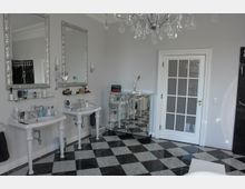 Luxuriöses Badezimmer mit schwarz-weiß kariertem Marmorboden, zwei Waschbecken mit silbergerahmten Spiegeln, einer Glastür mit weißen Rahmen und einem Kristallleuchter. Im Hintergrund steht ein verchromtes Sideboard.