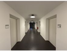 Ein heller, schlichter Flur mit weißen Wänden und dunklem Bodenbelag. Es gibt mehrere Türen auf beiden Seiten, beschriftet mit kleinen Schildern, und am Ende des Flurs befindet sich ein Gittertor.