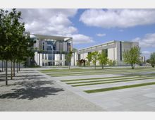 Modernes Regierungsgebäude mit großer Glasfassade und architektonisch markanten weißen Säulen; davor eine weitläufige, geometrisch gestaltete Grünfläche mit abwechselnden Streifen aus Rasen und Steinplatten. Einige Bäume spenden Schatten, und der Himmel ist teils bewölkt.