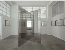 Minimalistischer Galerieraum mit weißen Wänden, gerahmten Zeichnungen an den Wänden und einem großen, geometrisch angeordneten Spiegelelement in der Mitte. Große Fenster mit Gitterrahmen lassen Tageslicht herein, und der Boden besteht aus grauem Beton.