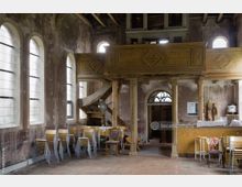 Innenraum eines historischen Gebäudes mit hohen, schmalen Rundbogenfenstern, einer hölzernen Empore mit verzierter Balustrade und einer abgenutzten Wendeltreppe. Im Vordergrund stehen gestapelte Holzstühle, während rechts eine improvisierte Theke mit Barhockern sichtbar ist.