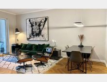 Wohnzimmer mit modernem Design, bestehend aus einem grünen Sofa, schwarzem Ledersessel mit Hocker, abstraktem Kunstwerk an der Wand, Glas-Kaffeetisch sowie einem Esstisch mit schwarzen Stühlen; Holzboden in Fischgrätmuster und großem Teppich. Im Hintergrund ein Fenster mit Zugang zu einem Balkon.