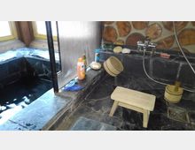 Traditionelles Badezimmer mit dunklem Steinboden, einem kleinen Holzschemel, Holz-Eimern und einer eingelassenen Badewanne aus dunklem Stein. Die Wand ist mit runden Holzelementen dekoriert, und Pflegeprodukte stehen auf einer Abstellfläche neben der Wanne.