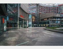 Platz in einem modernen Gebäude-Komplex mit glasüberdachtem Dach. Zu sehen sind mehrere Geschäfte wie ein Kino und ein Café, Fassaden mit großen Glasflächen sowie ein runder Gehweg mit gepflastertem Muster.