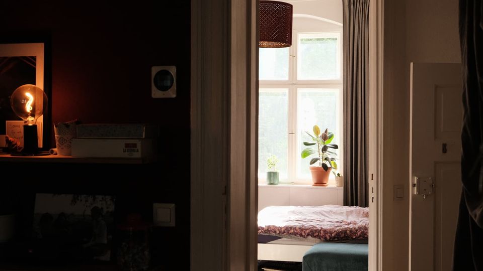 Blick durch einen Türrahmen in ein Schlafzimmer mit großem Sprossenfenster, grauen Vorhängen und Pflanzen auf der Fensterbank. Im Raum stehen ein Bett mit Decke sowie eine gepolsterte Sitzbank; links ist ein dunkler Flurbereich mit Regal und beleuchteter Lampe zu sehen.