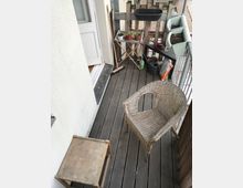 Ein kleiner Balkon mit einem Holzfußboden, darauf stehen ein Korbstuhl und ein kleiner Holztisch. An der Seite befinden sich Pflanzen in Töpfen und Pflanzkästen sowie Arbeitsgeräte wie ein Besen und eine Gießkanne.