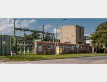 Freistehendes Umspannwerk mit einer Backsteinmauer im Vordergrund, die mit gemalten Fenstern und Ornamenten verziert ist. Im Hintergrund befinden sich grüne Metallstrukturen, Freileitungen sowie ein großes sandfarbenes Betongebäude; daneben Grünflächen und Bäume.