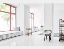 Heller, minimalistischer Innenraum mit weißen Wänden und Böden, großen Fenstern mit roten Rahmen und Sitzbänken mit Kissen. Im Raum befinden sich außerdem ein schwarzer Stuhl, ein Rollwagen, ein Regal mit Körben und ein kleiner Beistelltisch mit Blumen.