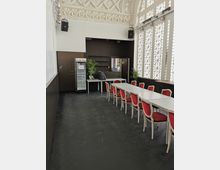 Ein Veranstaltungsraum mit hohen verzierten Decken, großen Fenstern mit geometrischen Mustern und ausgestatteten weißen Möbeln, darunter lange Tische und rote Stühle. Im Hintergrund stehen Pflanzen, ein Regal sowie ein Getränkekühlschrank.