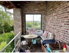 Gemütlicher Balkonbereich mit Ziegelwänden, einem Holzboden und einer Sitzbank mit bunten Kissen. Im Vordergrund steht ein runder Holztisch, umgeben von Pflanzen und mit Blick auf einen grünen Garten durch ein großes Fenster.