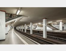 U-Bahn-Station mit mehreren Gleisen und Säulenreihen, weißen gefliesten Wänden und hell beleuchtetem Bahnsteig. Eine Treppe führt zu einer höheren Ebene, und einige Personen sind auf den Bahnsteigen zu sehen.