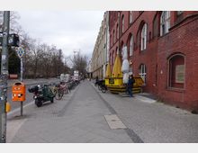 Straßenansicht mit Backsteingebäude auf der rechten Seite und einer langen Reihe geparkter Fahrräder. Auf dem breiten Gehweg sind eine orangefarbene Mülltonne, ein gelber Container mit zusammengefalteten Sonnenschirmen und ein Mann zu sehen, während links die Straße mit wenigen geparkten Fahrzeugen und kahlen Bäumen verläuft.