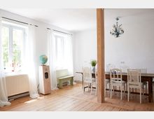 Heller Essbereich mit Holzboden, weißen Wänden und großen Fenstern mit transparenten Vorhängen. In der Mitte steht ein langer Holztisch mit weißen Stühlen, daneben eine Säule aus Holz und ein Podest mit einem Globus.