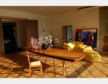 Ein heller Wohnraum mit Parkettboden, einem großen Holztisch, einer Vase mit rosa Blüten und einer gelben Polstersitzgruppe. Im Hintergrund sind Holztüren, eine offene Durchgangstür und ein farbenfrohes abstraktes Gemälde zu sehen.