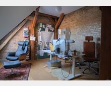 Ein Arbeitszimmer mit sichtbaren Backsteinwänden und Holzbalken. Im Raum befinden sich ein Schreibtisch mit Computer und Schreibtischlampe, ein lederner Bürostuhl, ein Sessel mit Fußhocker sowie ein Bücherregal aus gestapelten Büchern.