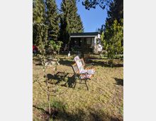 Garten mit Rasenfläche, Gartenbank im Vordergrund und blühendem jungen Baum; im Hintergrund steht ein kleines Haus mit überdachter Veranda, umgeben von hohen Nadelbäumen und Sträuchern.