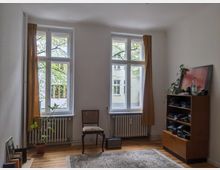 Ein minimalistisch eingerichtetes Zimmer mit zwei großen, weißen Holzrahmenfenstern, durch die Bäume und eine Hausfassade sichtbar sind. Der Raum hat weiße Wände, einen Holzboden, gelbe Vorhänge, einen Teppich, einen Stuhl, ein Regal mit Kleidung und Pflanzen sowie einige eingerahmte Bilder, die an der Wand lehnen.