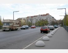 Straßenansicht einer Brücke in einer Stadt mit Wohnhäusern im Hintergrund. Auf der Straße parken ein Wohnmobil und mehrere Pkw; der Bürgersteig ist durch halbkugelförmige Poller abgegrenzt, und Laternen säumen die Straße.