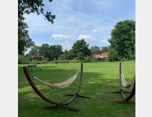 Zwei Hängematten mit Holzgestellen auf einer großen, grünen Wiese, umgeben von Bäumen. Im Hintergrund sind ein Baumhaus und ein Gebäude mit roten Ziegeldächern zu sehen.