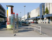Straßenansicht mit breitem Gehweg, einer Litfaßsäule mit Plakaten, einer Telefonzelle aus Glas und Edelstahl sowie parkenden Autos entlang eines langen, modernen Gebäudes mit großen, blauen Fensterelementen. Im Vordergrund ist eine Taube auf dem Gehweg zu sehen.