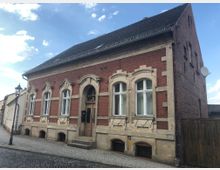 Ein zweigeschossiges Backsteinhaus mit verzierten Fensterrahmen und einer Holztür, gelegen an einer gepflasterten Straße. Das Gebäude hat ein steiles Schrägdach und steht neben einem Holzzaun; im Vordergrund befindet sich eine Straßenlaterne.