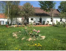 Ländlicher Garten mit einer Wiese, blühenden Blumenbeeten und mehreren Lehmbedeckten Töpfen vor einem weiß gestrichenen Haus mit Ziegeldach. Im Vordergrund stehen Tulpen und im Hintergrund sind Tische und Stühle auf einer Terrasse zu sehen, umgeben von Bäumen.