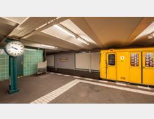 U-Bahn-Station mit grünen Wandfliesen, einer großen analogen Uhr auf einem Metallpfosten und einem gelben U-Bahn-Wagen der BVG auf der rechten Seite. An der Wand ist ein Schild mit der Aufschrift „Wittenbergplatz“ zu sehen.