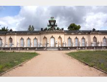 Barockes Bauwerk mit heller Fassade, großen Rundbogenfenstern und dekorativen Skulpturen, die entlang der gesamten Fassade stehen. Im Zentrum ist ein Portikus mit einer weißen Tür und einem Uhrturm mit grünem Kupferdach zu sehen, umgeben von einem symmetrischen Gartenweg.