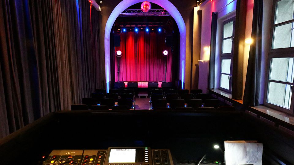 Blick aus dem Technikbereich in einen kleinen Theatersaal mit Sitzreihen und einer Bühne mit rotem Vorhang unter einem großen Rundbogen. Rechts sind hohe Fenster und Wandlampen zu sehen, die den Raum in farbigem Bühnenlicht beleuchten.