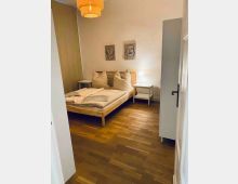 Schlichtes Schlafzimmer mit Holzbett, weißen Kissen und Bettwäsche auf dunklem Holzboden. An der weißen Wand hängen zwei abstrakte Bilder; daneben stehen zwei kleine Nachttische mit Lampe, und rechts ist ein hoher Kleiderschrank sowie eine offene Tür zu sehen.