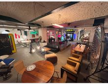 Ein loftartiger Raum mit gewölbter Ziegeldecke, eingerichtet mit unterschiedlichen Retro-Möbeln, Tischen und Stühlen. Im Hintergrund sind Neonlichter, Schilder mit "BERLIN" und "seit", sowie moderne Lampen und Dekorationen sichtbar.