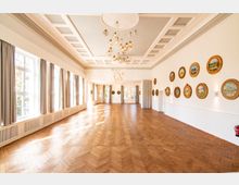 Heller Saal mit Parkettboden, großen Fenstern mit Vorhängen auf der linken Seite und dekorativen, runden Bildern an der weißen rechten Wand. Eine stilvolle Decke mit integrierten, hängenden Lampen sowie ein großzügiger, lichtdurchfluteter Raumcharakter definieren die Architektur.