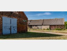 Hofbereich mit alten Wirtschaftsgebäuden: links eine Backsteinfassade mit großem grauem Metalltor und Graffiti, rechts ein langes Stall- oder Scheunengebäude mit Wellblechdach und Holztoren. Dazwischen verlaufen ein Grasstreifen und ein gepflasterter Weg mit einer kleinen Wegtafel und Laterne unter blauem Himmel.
