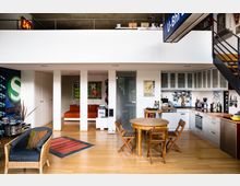 Offener Wohnraum in einem modernen Loft mit Holzboden. Zu sehen sind eine Küche mit weißen Schränken und Edelstahlgeräten, ein Essbereich mit rundem Holztisch und Stühlen, dekorative Poster an den Wänden und eine Treppe, die zu einer offenen Galerie führt.