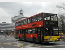 Ein roter Doppeldeckerbus der Linie 104 mit gelbem Frontbereich fährt auf einer belebten Straße vorbei, im Hintergrund stehen moderne Bürogebäude, darunter ein Gebäude mit RBB-Logo. Straßenmarkierungen und Baustellenabsperrungen sind ebenfalls sichtbar.